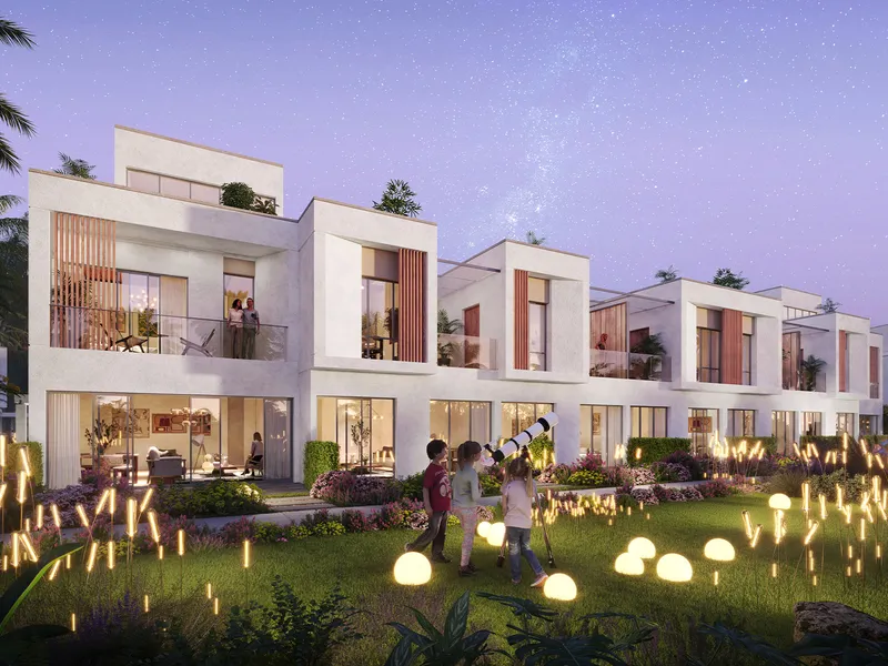 Row of white three-storey DAMAC Riverside townhouses at dusk, with a family on the lit lawn in front