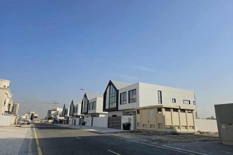 Site photograph of a finished villa street at Nad Al Sheba Gardens, gabled roofs and gated driveways, with a tower crane working on a later phase behind