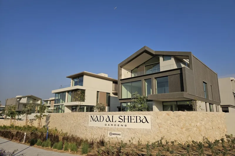 Site photograph of finished gabled villas behind the stone Nad Al Sheba Gardens entrance wall carrying the Meraas mark