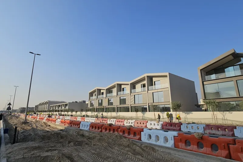 Site photograph of a completed townhouse terrace and the community building at Nad Al Sheba Gardens, with road construction and barriers along the frontage