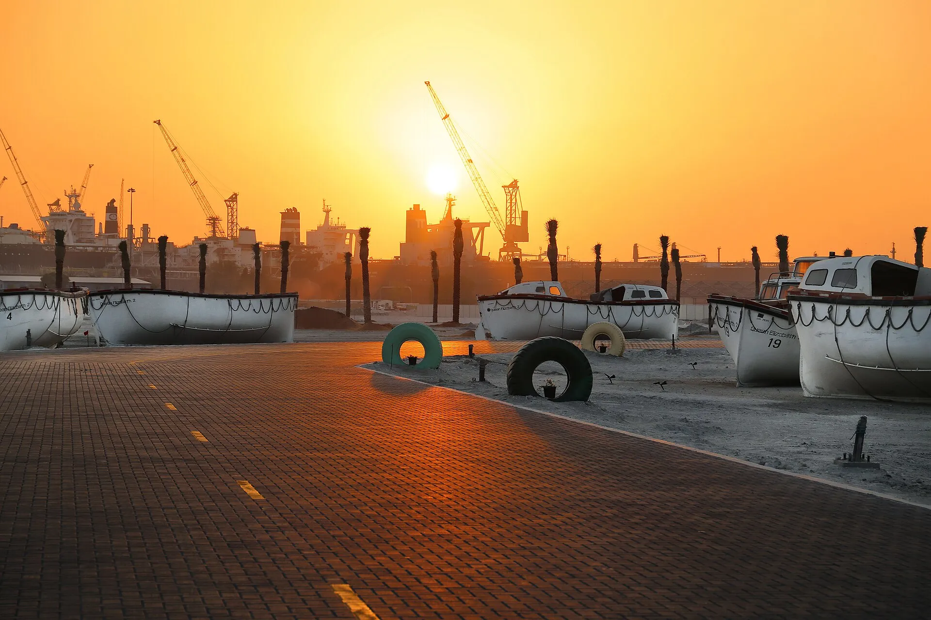 Lifeboats marked Queen Elizabeth 2 on the quay at Port Rashid at sunset, with cargo cranes and ships silhouetted behind