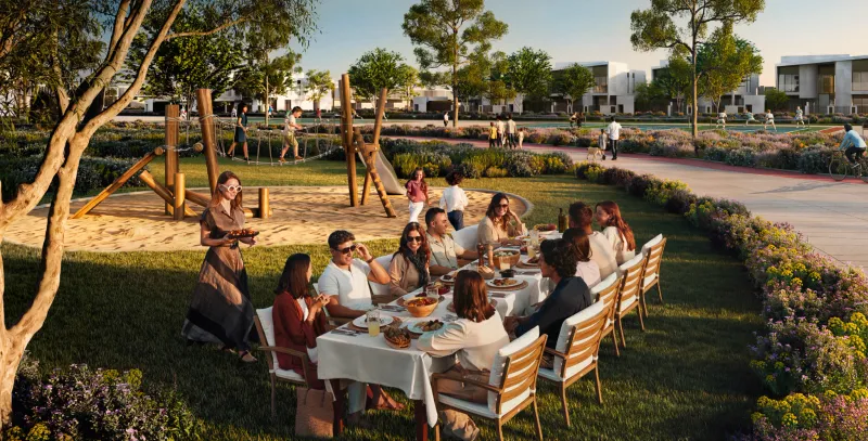 Render of a long dining table laid on a lawn in the Talay community park, with a rope-climbing playground, flowering planting, a cycling and jogging path, and rows of white flat-roofed houses behind
