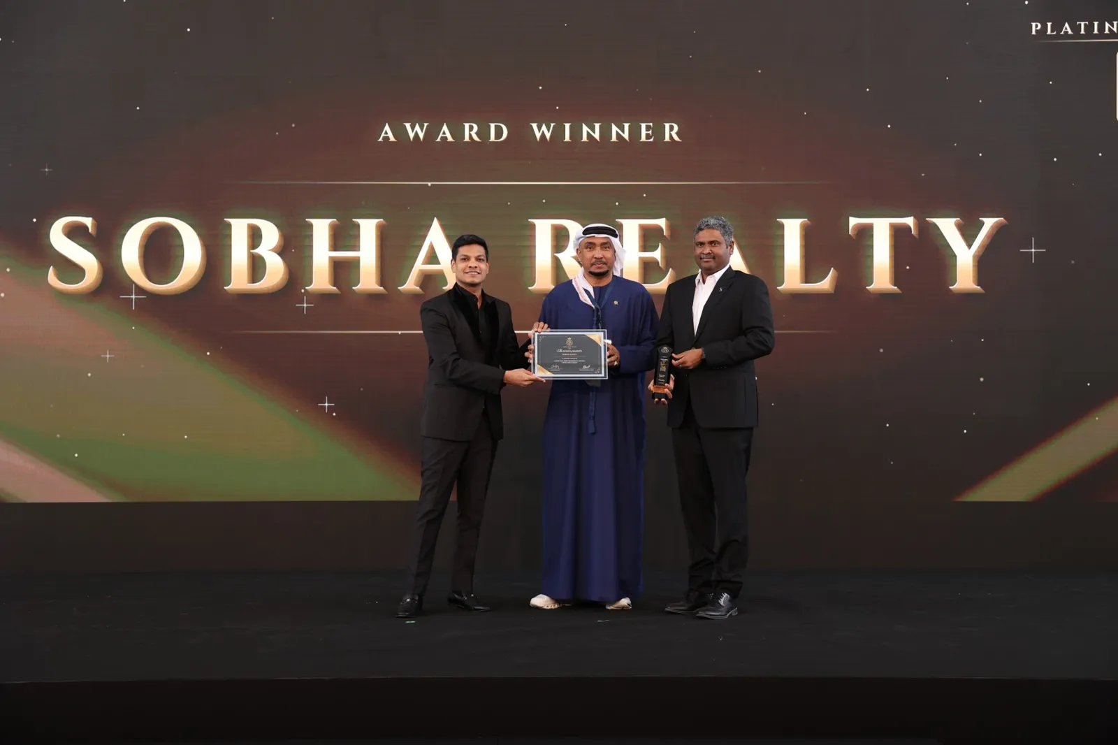 Two Sobha Realty executives receive a certificate and trophy on stage beneath a screen reading Award Winner, Sobha Realty