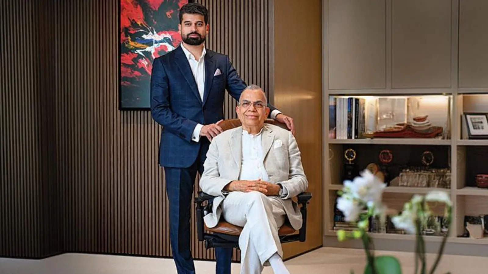 Two men in a wood-panelled office: one in a navy suit stands behind another seated in a light linen suit